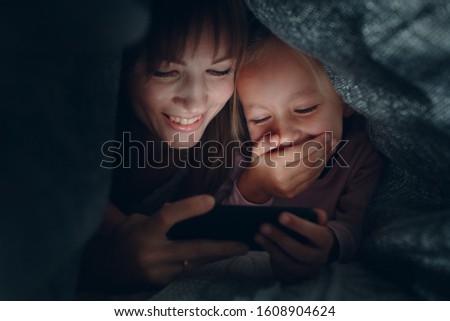Mother with a little daughter watching content on  smartphone in the dark under the blanket covers.