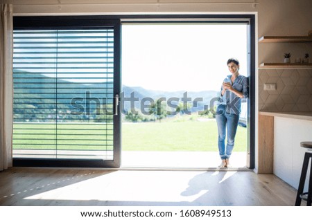 Front view of young woman with coffee standing by patio door at home. Royalty-Free Stock Photo #1608949513