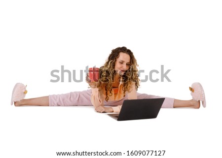 Young attractive woman in pajamas and slippers is sitting in the splits drinking morning coffee and working at a laptop isolated on a white background
