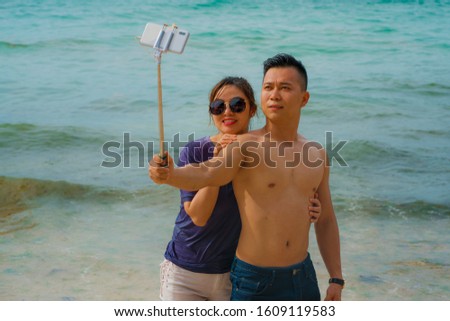 Summer holidays lifestyle portrait of young happy and playful Asian Chinese couple enjoying at the beach taking stick selfie photo with hand phone enjoying the sea together