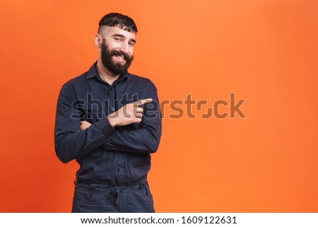 Image closeup of brunette man with nose jewelry wearing black shirt pointing finger aside at copyspace isolated over orange background