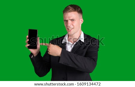 Business man in a jacket points a finger at a smartphone with a blank screen, isolated on a green background. Concept for advertising mobile applications.