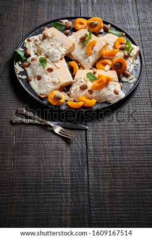 Yogurt ice cream of apricot and almond served with fresh summer apricots, almonds and almond slices decorated with mint on a black plate with dessert cutlery on a dark wooden table, vertical view