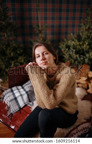 brunette girl sitting on a plaid with a christmas wreath and wood with a burner on a background of Christmas trees and a retro checkered pattern. Woman in a beige sweater in a cozy New Year's interior