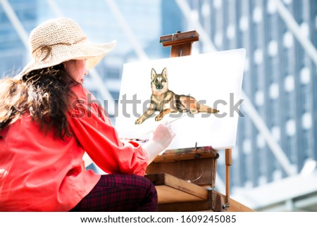Woman sitting and painting the dog her loves on canvas on a wooden easel with a brush at the street of Bangkok.Lifestyle of painter are painting her lovely dog.Street artists in holiday and hobby.