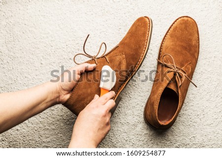 Hands cleaning men's camel suede desert shoes (boots) with a brush. Footwear maintenance captured from above (top view). Grey concrete background.  Royalty-Free Stock Photo #1609254877