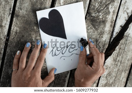Valentine's day card - black paper heart and white blank with the inscription charcoal - love, on a wooden background. Human, woman, hands writes. Copy space. Flat lay. Horizontal orientation.