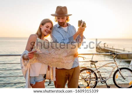 attractive happy couple traveling in summer on bicycles, man and woman with blond hair boho hipster style fashion having fun together, looking in map sightseeing taking photos on camera