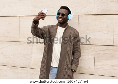 Portrait stylish smiling african man taking selfie picture by smartphone in wireless headphones listening to music wearing brown knitted cardigan, sunglasses on city street over wall background
