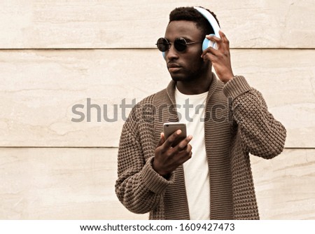 Portrait stylish african man with phone in wireless headphones listening to music wearing brown knitted cardigan and sunglasses on city street over brick wall background