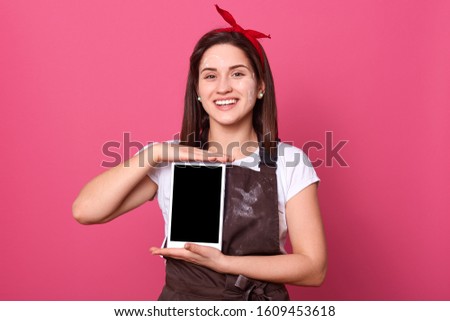 Picture of positive energetic housewife with flour on face and apron, wearing white t shirt and red headband, holding tablet with both hands, smiling sincerely, having pleasant facial expression.
