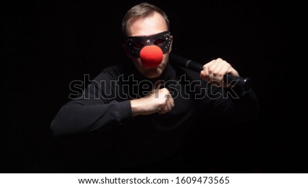 man in a leather mask with a red clown nose waving a bat on a black background.