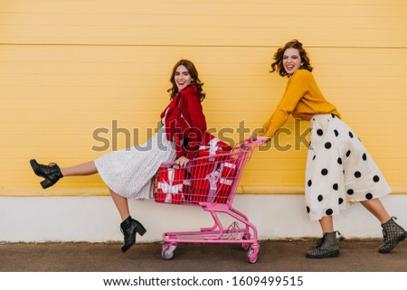 Jocund friends enjoying weekend shopping. Outdoor portrait of two girls with red present boxes.