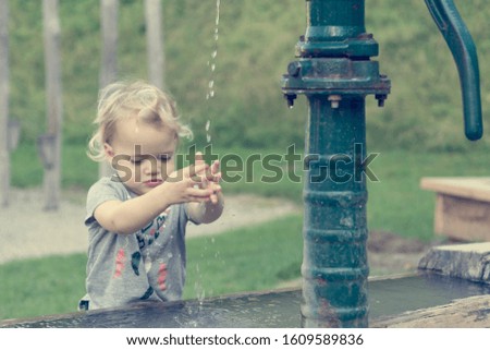 Cute blonde girl washing hands using hand water pump in local park.