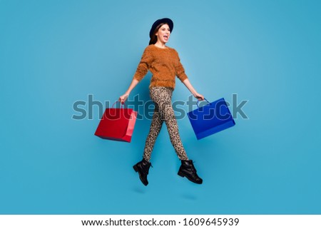 Full length photo of cheerful funky girl enjoy autumn spring holiday shopping  hold beautiful bags go jump walk wear brown pullover retro trousers headwear cap isolated blue color background