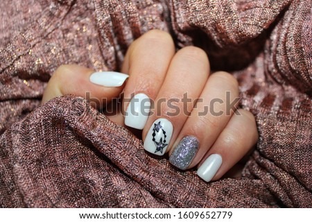 One female hand with white manicure with black and silver stars on beige background. Stylish fashionable woman's manicure. Nail polish. Artistic manicure. Modern style. Winter manicure.