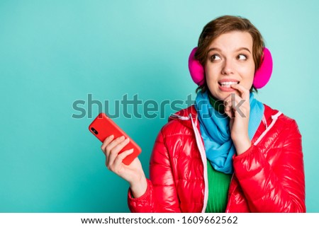 Photo of pretty beautiful funny lady hold telephone looking interested empty space biting finger wear red coat scarf pink ear covers green jumper isolated teal color background