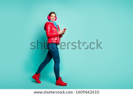 Full length profile photo of funny lady walking down street chatting telephone good mood wear casual red coat scarf pink ear covers pants shoes isolated teal color background