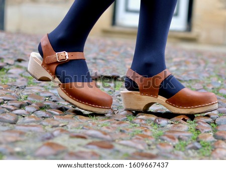 A close up of a woman's legs walking with beautiful leather and wood clog shoes on a cobblestone cobble street Royalty-Free Stock Photo #1609667437