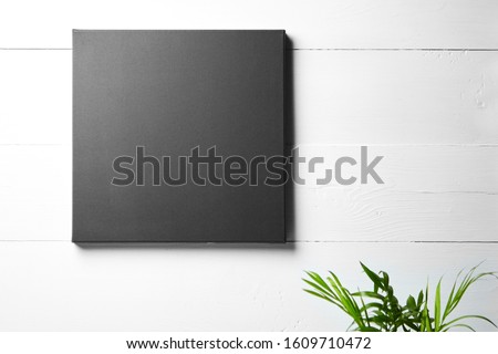 A black cotton canvas and leaves of green plant  on white wooden background. Stretched clean canvas hanging on wall. Mockup, front view