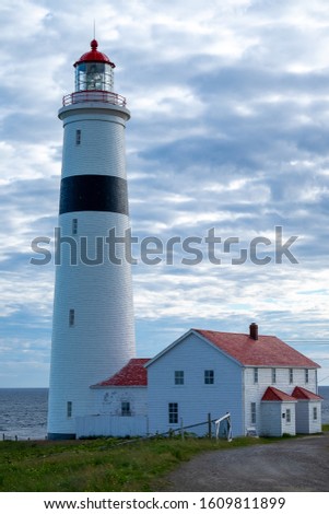 A white round style lighthouse with a red stripe adjoined to a wooden building with a red roof. The lighthouse is at the edge of a cliff with the ocean in the background. The sky is cloudy and bright.