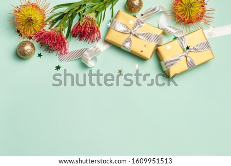 Gifts that are wrapped in gold paper with white ribbon surrounded by glitter and Australian native red and yellow waratahs, proteas on a green background. Space for copy.