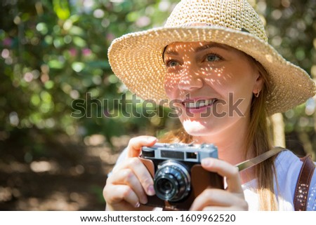 Close-up portrait of a beautiful woman in straw hat travelig in tropic forest, taking photos on retro camera. Light shadows through the cut-out detailed brim on face. Tourist with backpack. 