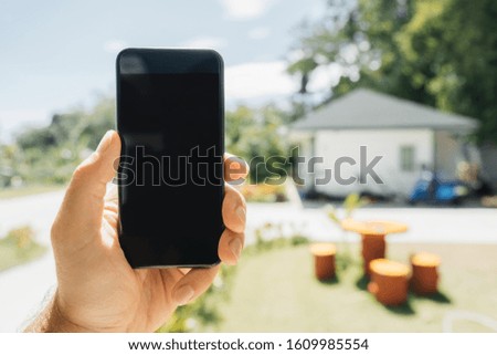 The smartphone screen is black. A hand holds a mobile phone with a view of a residential building in the summer, sunny.