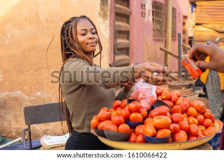 Young black woman arranging her tomatoes and pepper in the market and also receiving a card from someone to make payment