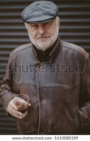respectable man and delicious alcohol. An elderly man with a gray beard in a leather jacket and cap holds in his hand a glass of whiskey
