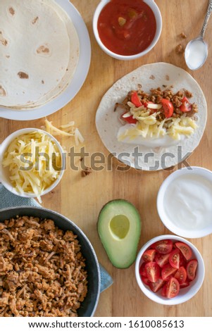 overhead flat lay of tacos for dinner with ground mince beef cheese hot sauce avocado tomato and sour cream on warm tortillas