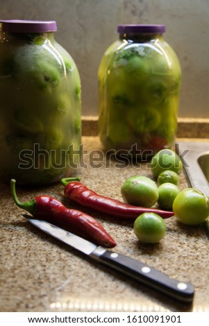 Still life with red pepper. Two red hot peppers and green tomatoes lie on a granite countertop. In the background are two glass jars with green tomatoes.
