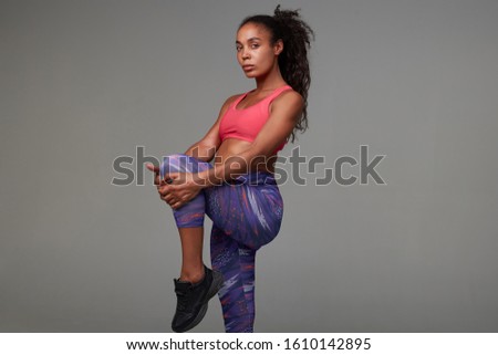 Studio photo of young sporty dark skinned woman wearing her curly long brown hair in ponytail hairstyle while stretching her muscules after hard training, isolated over grey background