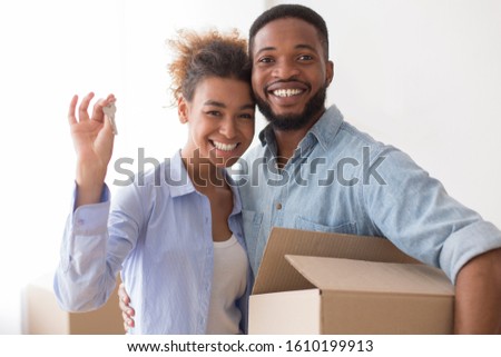 Own Home. Young Afro Spouses Showing New House Key Hugging And Holding Moving Box Standing Indoor.