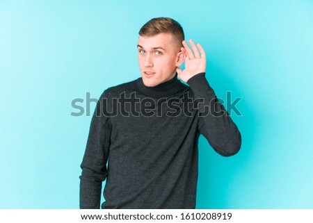 Young caucasian man on a blue background trying to listening a gossip.