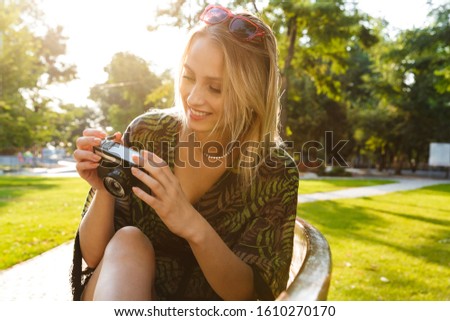 Beautiful happy young blonde woman sitting on a bench at the city park, holding photo camera