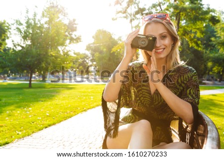 Beautiful happy young blonde woman sitting on a bench at the city park, holding photo camera
