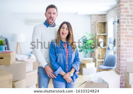 Young beautiful couple standing at new home around cardboard boxes sticking tongue out happy with funny expression. Emotion concept.