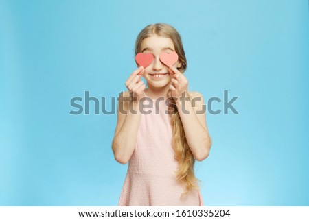 Isolated little girl covers her eyes with paper hearts on a blue background. Isolated half-length portrait. Little girl and paper hearts.
