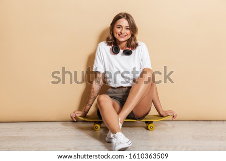 Portrait of a smiling schoolgirl sitting on a skateboard isolated over beige background, relaxing