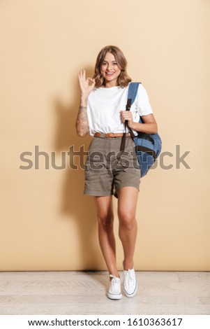 Image of happy teen girl carrying backpack smiling and showing ok sign isolated over beige background