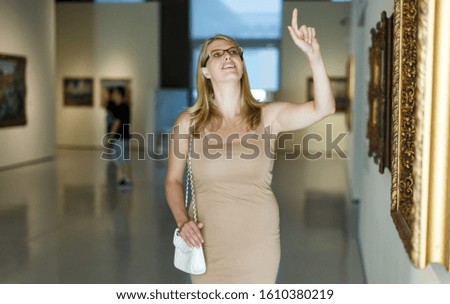Positive woman wearing glasses standing in hall of picture gallery and pointing to invisible screen