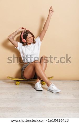 Image of beautiful teen girl in headphones rejoicing while sitting on skateboard isolated over beige background