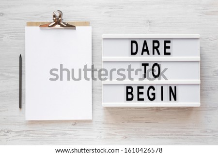'Dare to begin' words on a lightbox, clipboard with blank sheet of paper on a white wooden background, top view. Overhead, from above, flat lay. Copy space.