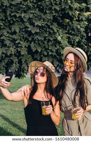 Two pretty girls take selfie in the summer outdoors at sunset. Girlfriends have fun, laugh, smile and take pictures. Closeup portrait of two young women in straw hats.