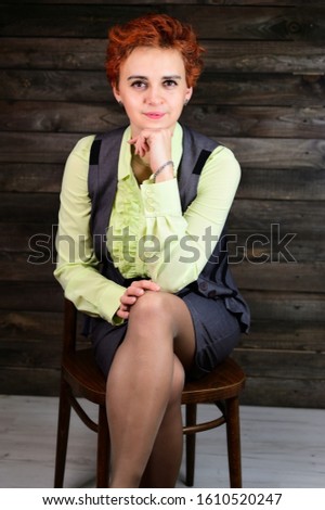 A woman sits on a chair right in front of the camera in various poses. Art portrait of a pretty business model with short red hair in a business suit on a wooden background in the interior.
