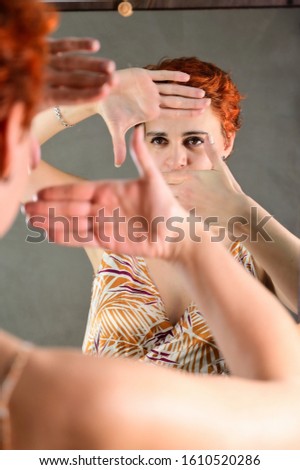 A woman poses in front of a mirror in front of the camera in various poses. Art portrait of a pretty model with short red hair in light overalls on a gray alternative background.