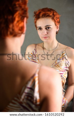 A woman poses in front of a mirror in front of the camera in various poses. Art portrait of a pretty model with short red hair in light overalls on a gray alternative background.