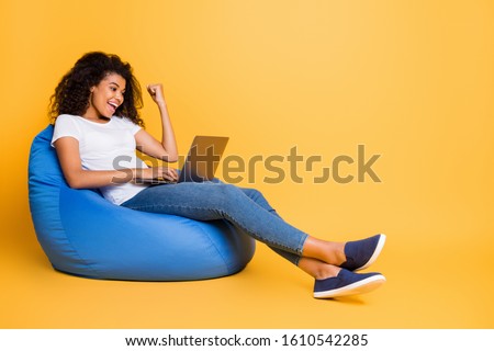 Portrait of nice attractive lovely cheerful cheery glad satisfied wavy-haired girl sitting in bag chair using laptop celebrating isolated over bright vivid shine vibrant yellow color background