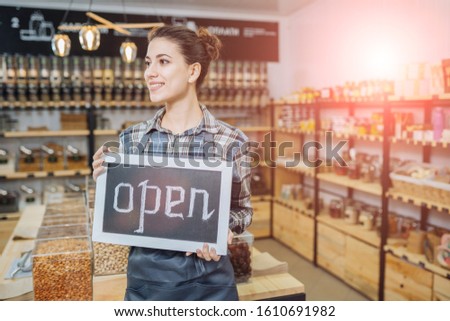Female business owner holding OPEN sign in grocery over zero waste shop interior. Sun glare effect. Conscious minimalism vegan lifestyle concept.